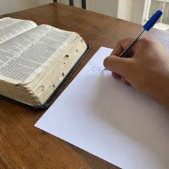Mão escrevendo uma frase no alto da folha, ao lado da Bíblia